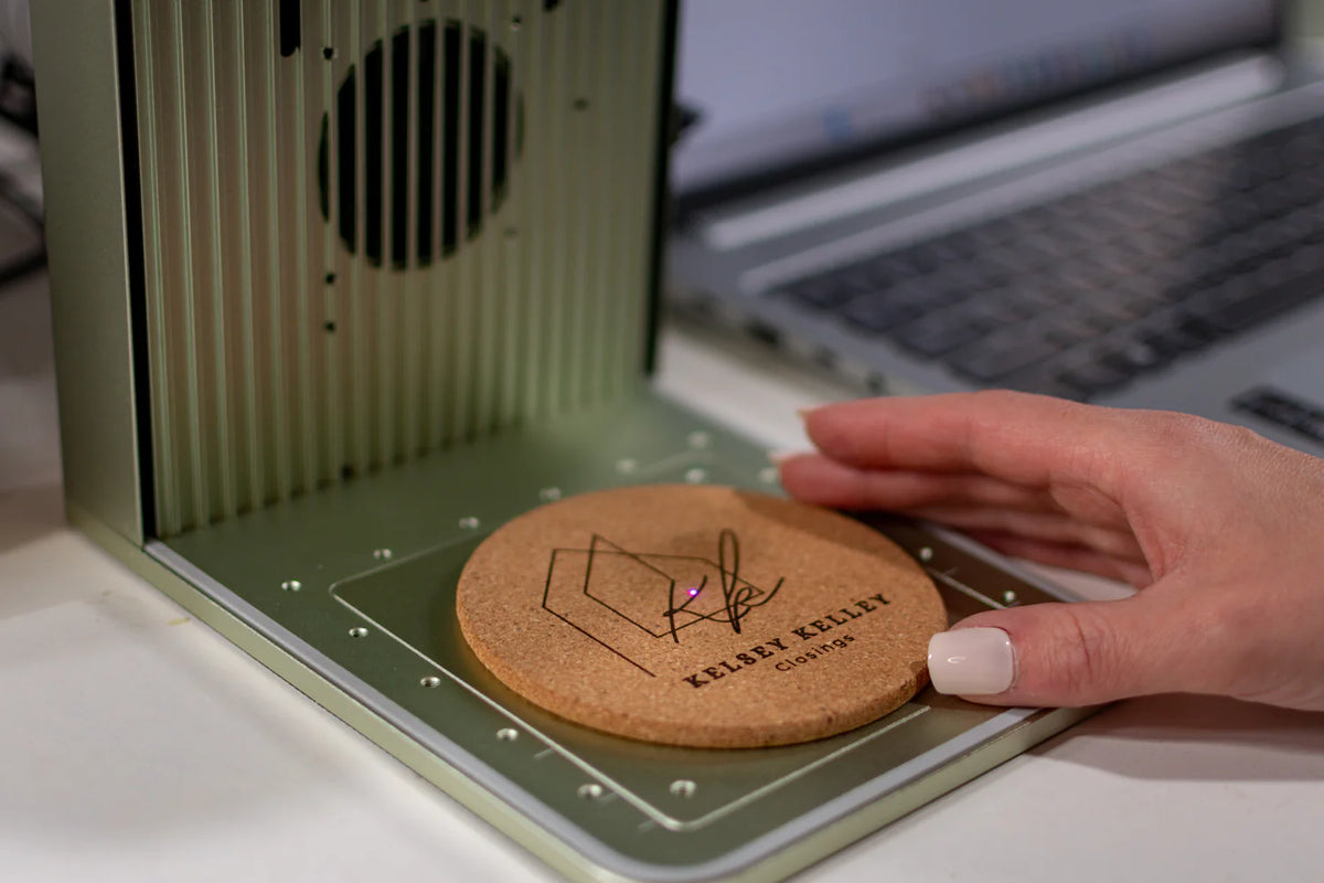 A close-up of a custom engraved cork coaster being held by a hand next to a mint green laser engraving machine, with a blurred laptop in the background.
